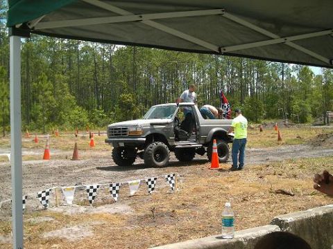 1986 Ford Bronco - A modified pickup truck sits on a dirt track, with people gathered around it and a checkered flag line in the foreground.