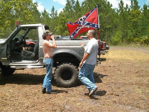 1986 Ford Bronco - Two men stand beside a lifted pickup truck flying a Confederate flag, with another vehicle visible behind it.