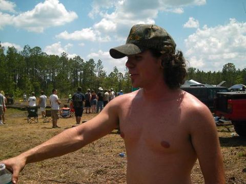 1986 Ford Bronco - A man in a camouflage cap stands shirtless in a field with a crowd and vehicles gathered under a cloudy sky.