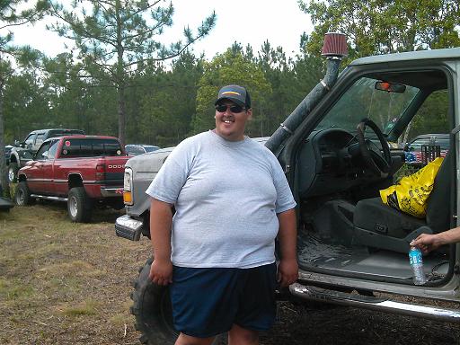 Man in t-shirt and shorts stands by a lifted pickup truck with snorkel; red Dodge pickup in background