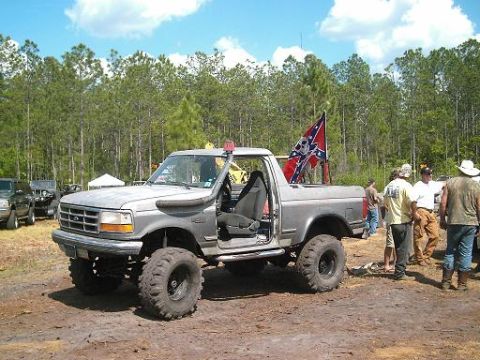 1986 Ford Bronco - A modified Ford Bronco with a Confederate flag is parked in a dirt lot with trees in the background.