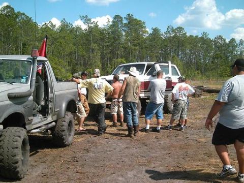 1986 Ford Bronco - A group of people stand around two modified off-road vehicles in a dirt lot surrounded by trees.