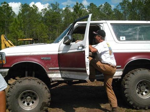 1986 Ford Bronco - A man climbs into a lifted white and maroon Ford Bronco with off-road tires.