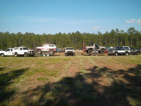 1986 Ford Bronco - A group of modified trucks and SUVs are gathered in an open field under a clear blue sky.