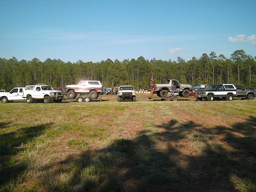 Several lifted trucks and SUVs parked in a grassy field with a line of trees in the background. One white Bronco is on a trailer