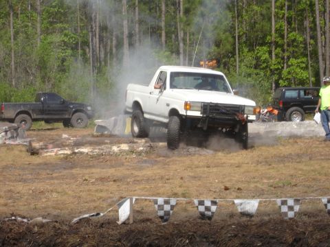 1989 Ford Bronco - A white pickup truck navigates an obstacle course, with other vehicles visible in the background.