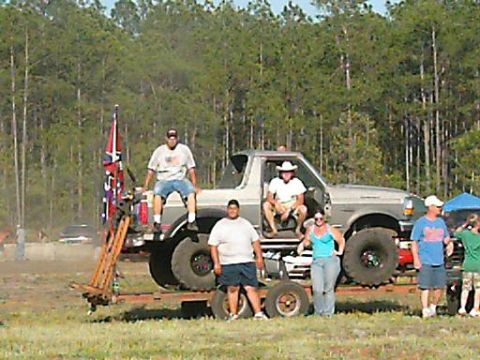 1986 Ford Bronco - Several people stand around a large, lifted Ford truck on a trailer in a grassy field.
