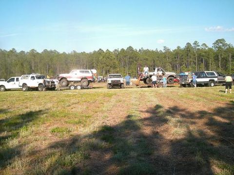 1986 Ford Bronco - A group of off-road vehicles are gathered in a grassy field with a line of trees in the background.