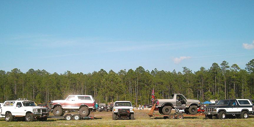 Several modified trucks and SUVs parked in a field with trees in the background. One white Bronco is on a trailer