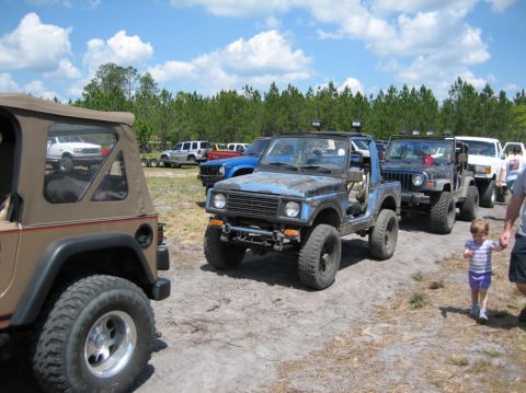 1989 Ford Bronco - Several off-road vehicles are lined up on a dirt path under a partly cloudy sky.