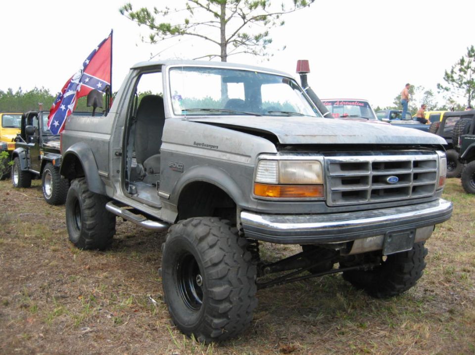 Lifted Ford Bronco with large tires, driver's door open. Another Jeep is parked behind it