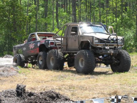 1989 Ford Bronco - Two custom monster trucks are positioned in a field, with one truck appearing to be towed or connected to the other.