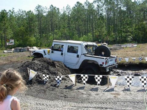 1989 Ford Bronco - A white Hummer H1 is stuck in a mud pit during an off-road event, with a young girl observing.