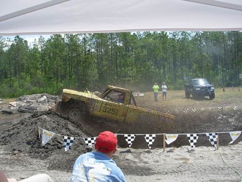 1989 Ford Bronco - A yellow pickup truck navigates a mud obstacle course with spectators watching.