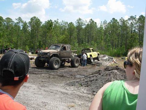 1989 Ford Bronco - Modified off-road trucks are parked in a dirt area with trees and debris around them.