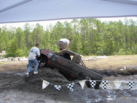 1989 Ford Bronco - A modified truck is stuck in a mud pit during an event, with two people attempting to push it free.