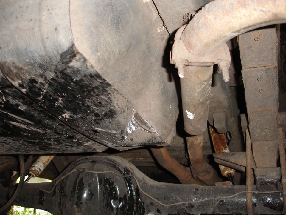 Underside view of a vehicle's fuel tank and exhaust system, showing rust and wear