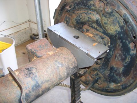 1990 Ford Bronco - A bare, heavily rusted rear axle housing rests on a jack stand in a garage while a new bare-steel leaf-spring perch is test-fitted on the t…
