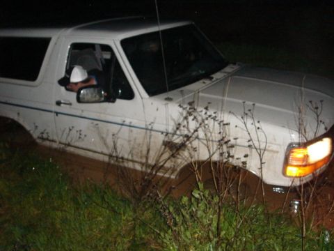 1994 Ford Bronco - A white 1994 Ford Bronco is stuck in the mud at night, with its headlights on.