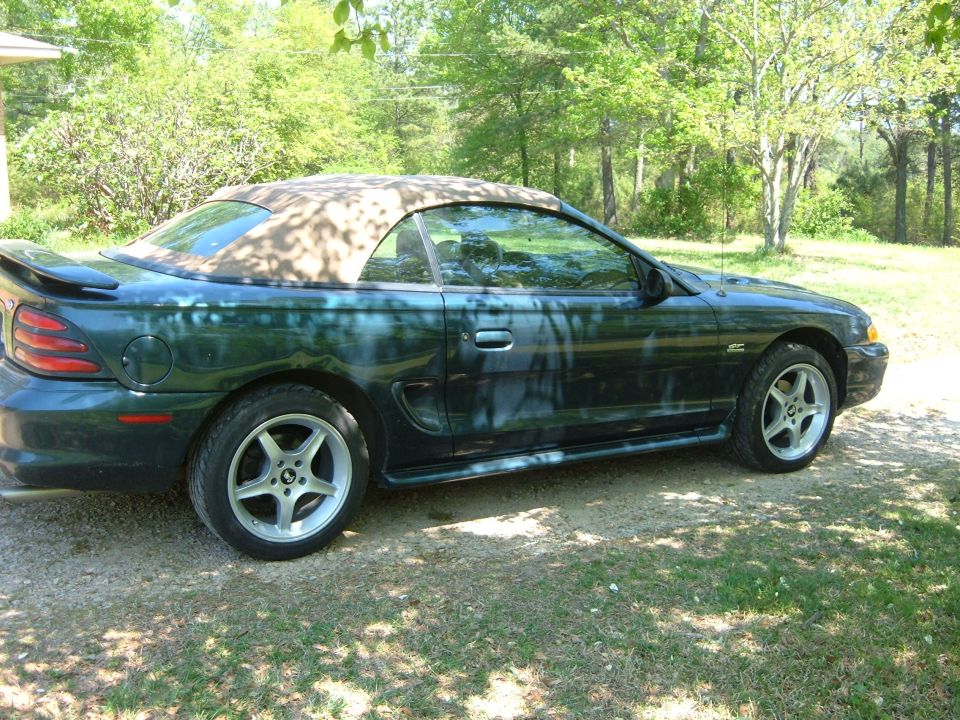 Dark green Ford Mustang convertible with tan top, side view, parked on gravel and grass