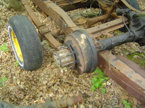 1979 Ford Bronco - Close-up of a rusty truck frame with rear axle and wheel, resting on the ground.
