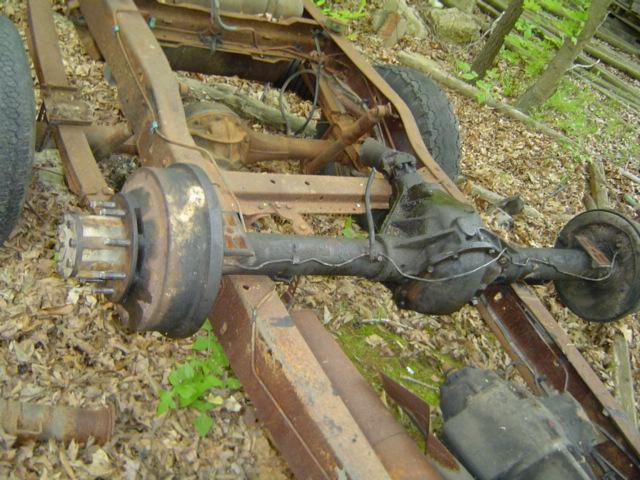 rusted vehicle frame with rear axle and drum brakes, sitting on the ground outdoors