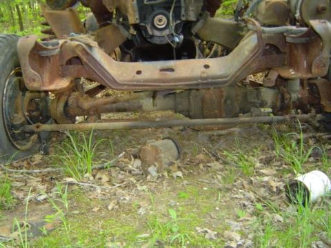 1979 Ford Bronco - The heavily rusted front chassis and engine components of a vehicle are visible, with grass and debris on the ground.