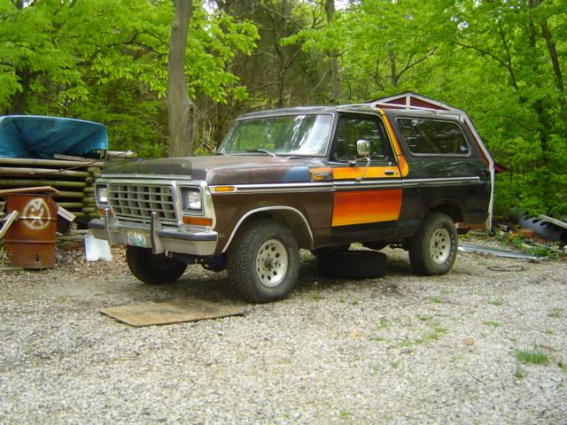 Brown and black 1978 Ford Bronco with orange and yellow stripe, parked on gravel