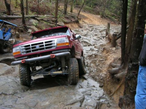1995 Ford Bronco - This red SUV is tackling a challenging off-road trail with large rocks and mud.