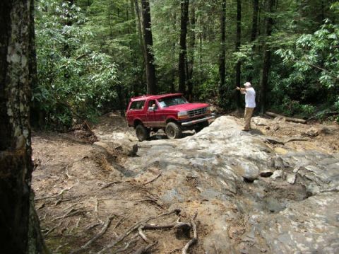 1995 Ford Bronco - A person stands on a rock, pointing towards a red SUV as it drives on a challenging off-road path.