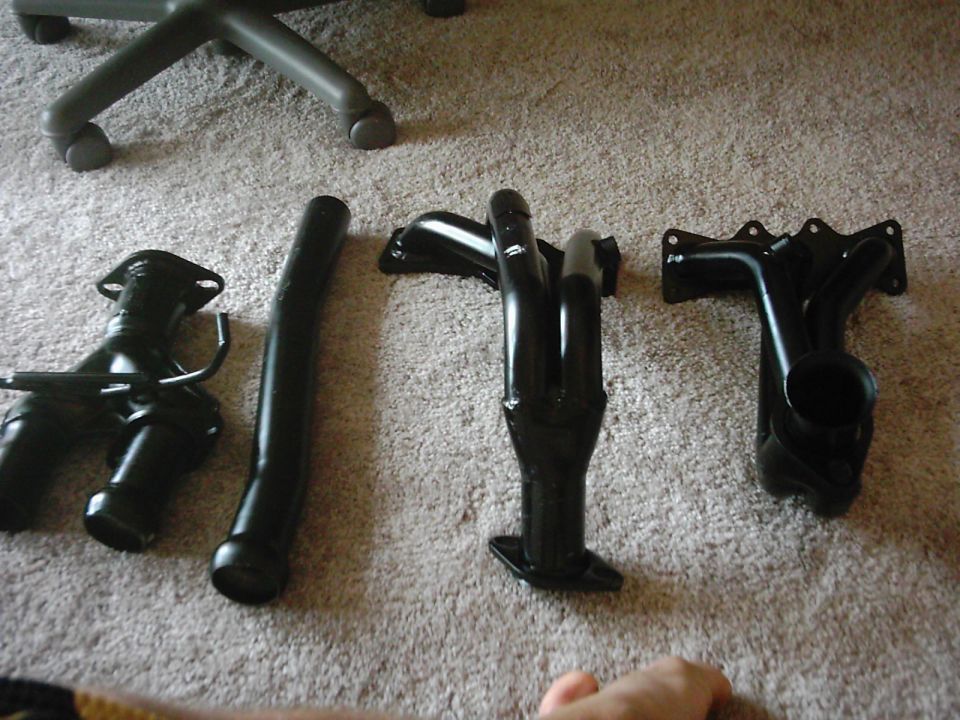 Four black-painted exhaust headers and tubes laid out on beige carpet