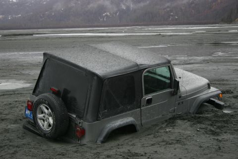 2003 Jeep TJ - A silver Jeep TJ is partially submerged in mud near a body of water with a forested hill in the background.