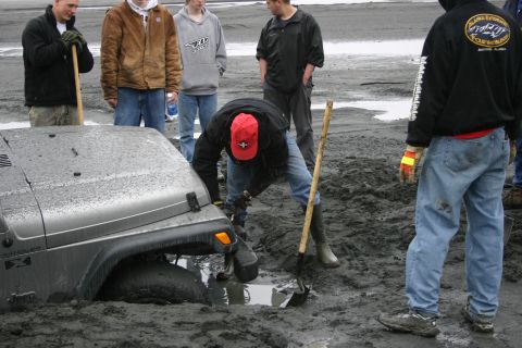 2003 Jeep TJ - A group of people attempt to dig a silver Jeep Wrangler out of thick mud.