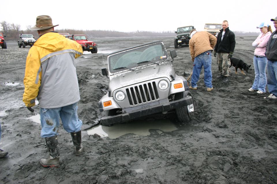 Silver 2003 Jeep Wrangler stuck in mud, front view. People and other vehicles nearby