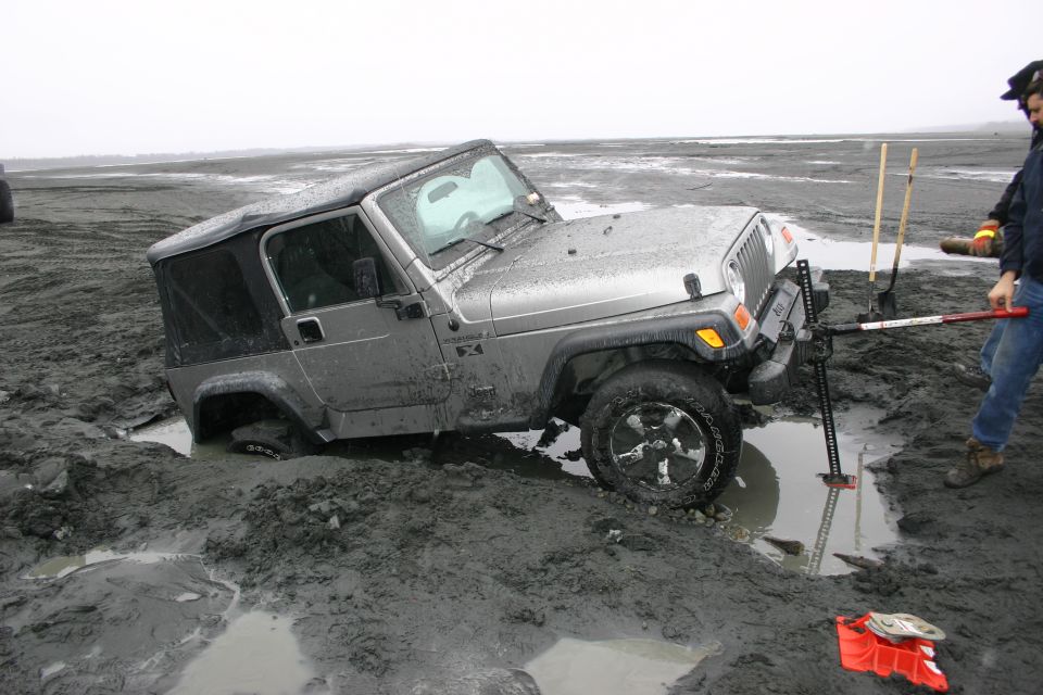 Silver Jeep TJ stuck in mud with people trying to free it using a jack and shovels