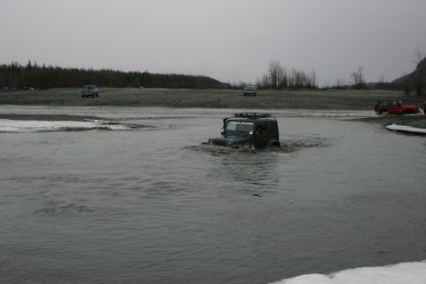 2003 Jeep TJ - A dark Jeep Wrangler navigates through shallow water, with other vehicles visible in the background.