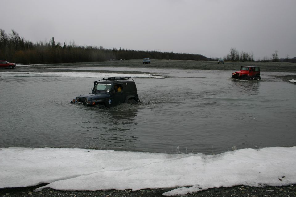 Dark green Jeep TJ driving through shallow water with a roof rack. Red Jeep visible in background