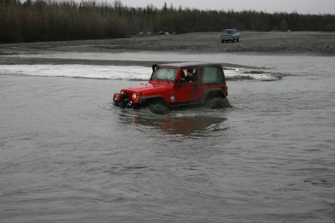 2003 Jeep TJ - A red Jeep TJ navigates through a shallow body of water, kicking up mud and spray.