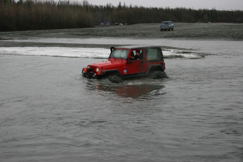 Red Jeep TJ driving through shallow water with a snorkel. Another blue SUV is visible in the background