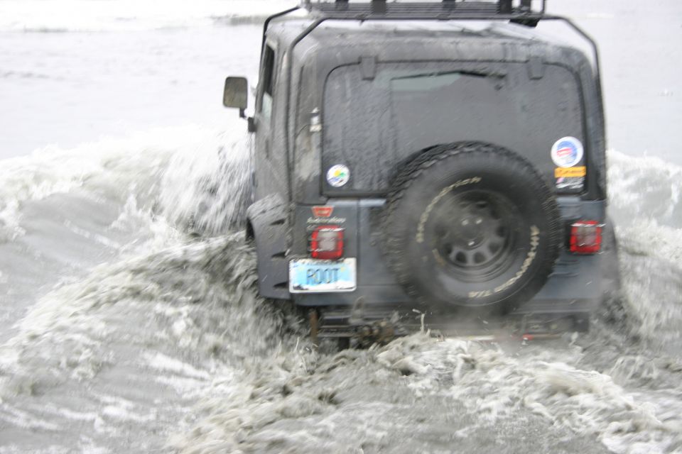 Dark gray 2003 Jeep TJ driving through water, rear view, spare tire on back, license plate reads ROOT