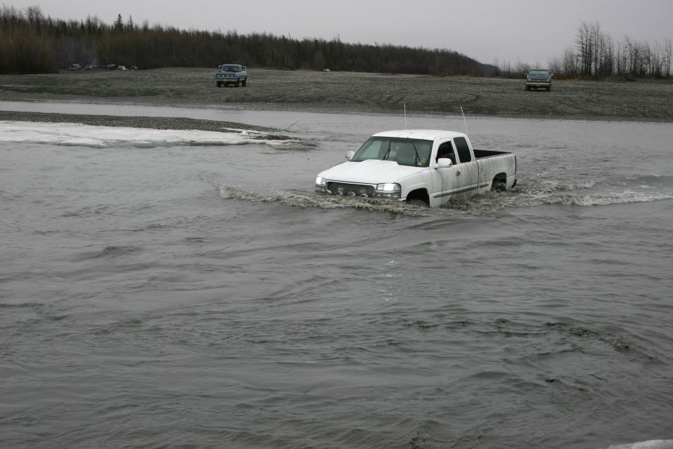 White GMC Sierra pickup truck driving through a shallow river, another truck in background