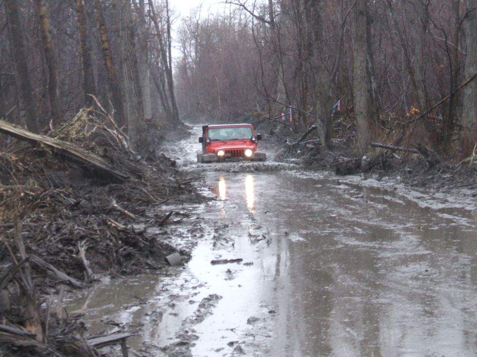 Red Jeep with headlights on driving toward camera on a flooded muddy trail in bare woods