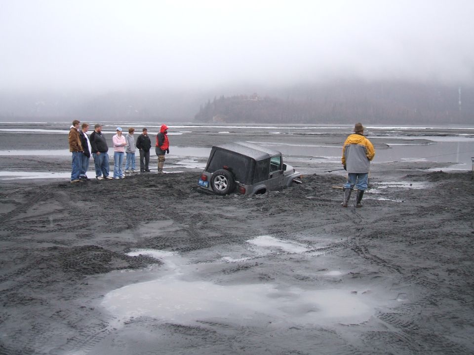 Jeep sunk to its body in gray tidal mudflat with onlookers and a man in waders