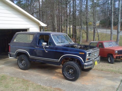 1984 Ford Bronco - A blue Ford Bronco is parked in front of a garage, with its hood up, next to an orange pickup truck.