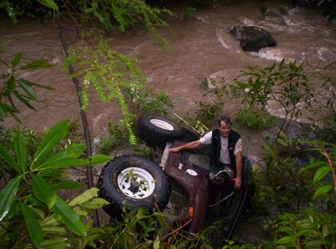 1995 Ford Bronco - Overhead view of a dark red Jeep lying on its side in a fast-moving muddy creek, showing its mud-terrain radial tires on white spoke wheels…