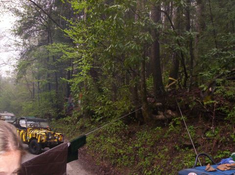 1995 Ford Bronco - Trail scene on a wet gravel forest road where a yellow tiger-striped open-top 4x4 with a roll bar and knobby tires sits behind a winch cabl…