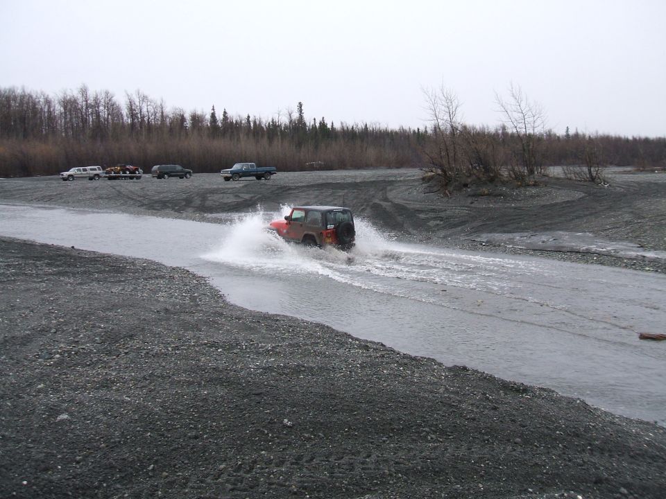 Orange Jeep splashing through a shallow river crossing with parked trucks on the far gravel bank