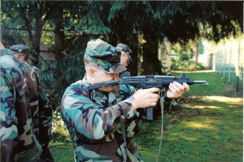 2003 Jeep TJ - A soldier practices aiming a submachine gun outdoors, with trees and a fence visible.