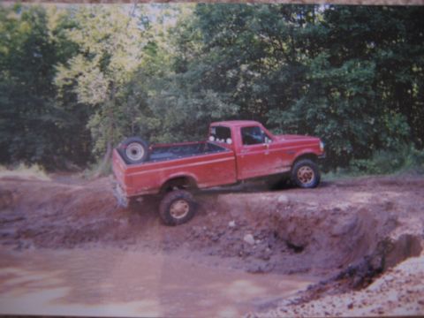 1977 Ford F-150 - A red pickup truck navigates a challenging off-road course, with its rear wheels on a steep, muddy embankment.