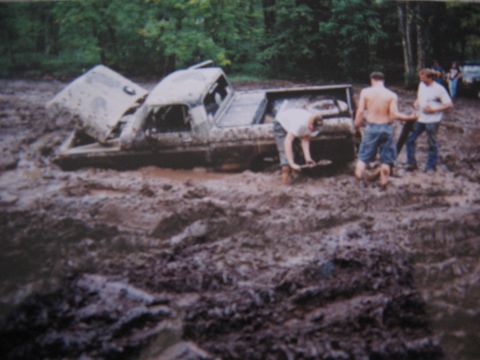 1977 Ford F-150 - A 1977 Ford F-150 is deeply stuck in mud, with its hood ajar and people attempting to free it.